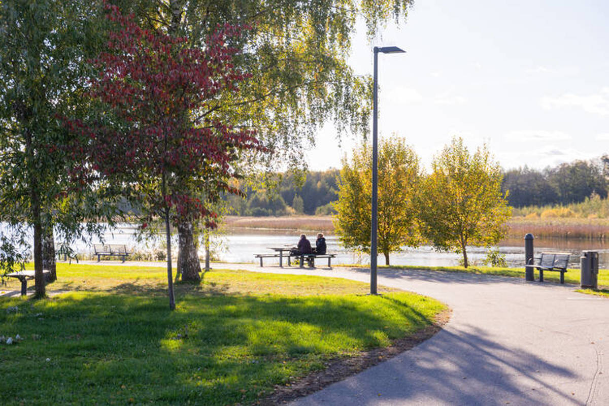 Valokuvanäkymä Järvenpään Rantapuistostasta, jossa kaksi henkilö istuu penkillä katsomassa järvelle päin